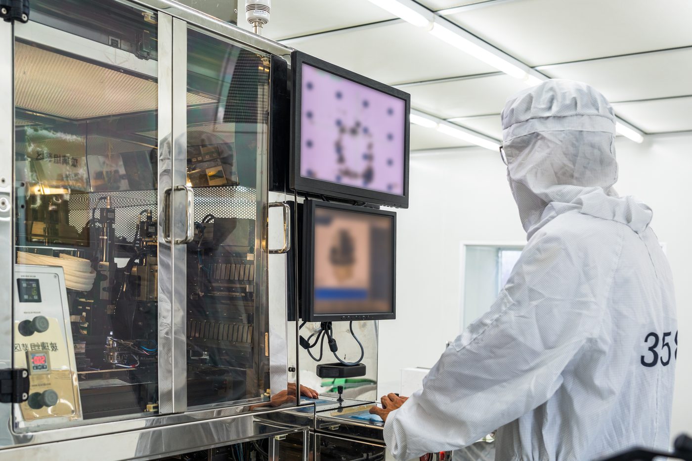 Technician in a cleanroom suit at a process tool, watching its two displays